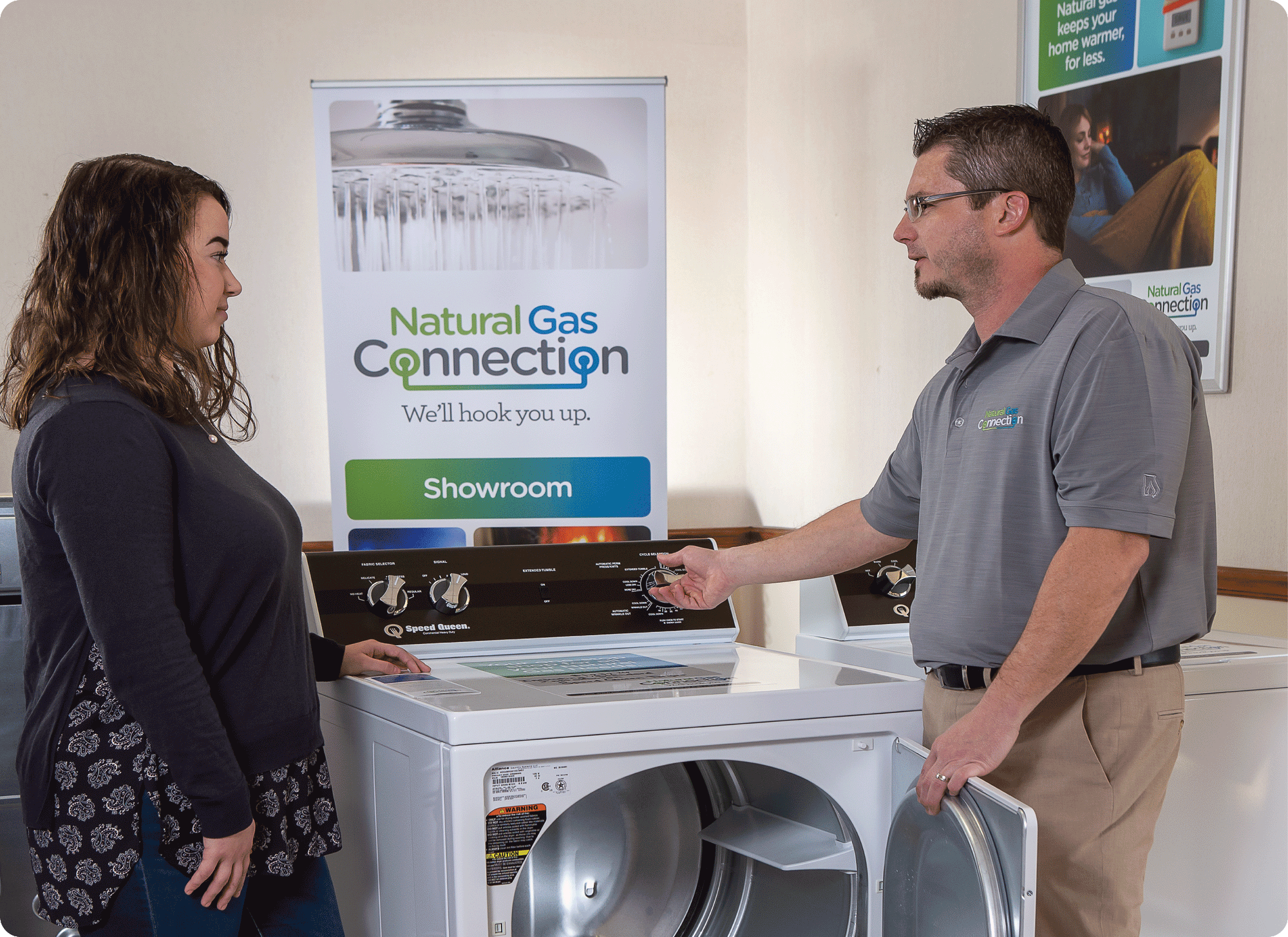 Sales associate discussing Speed Queen gas dryer with customer in appliance showroom, surrounded by business posters on Natural Gas Connection