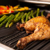 Close-up of BroilKing Crown 420 grill with cast iron grids cooking chicken legs, asparagus, and peppers